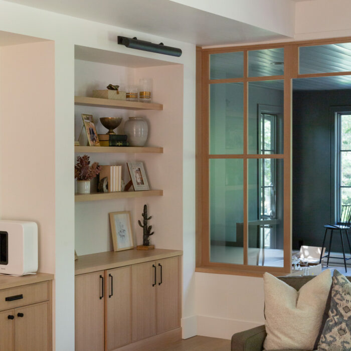 Built-in wooden shelves and cabinets with decorative items, next to a large window and a brown couch with patterned pillows in a modern living room.