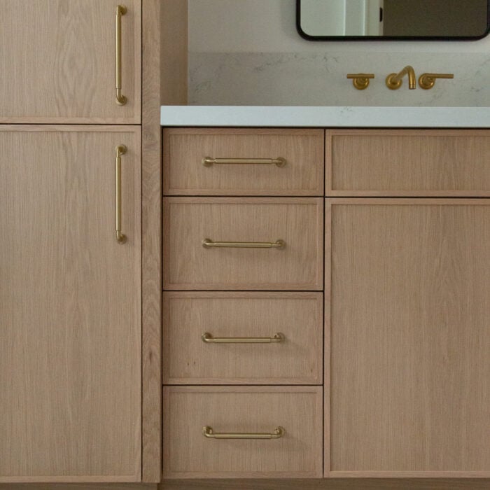 Light wood bathroom vanity with gold handles, white countertop, brass faucet, and a rectangular mirror above. The floor has diagonal, light-colored tiles.