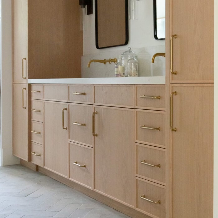 Light wood bathroom vanity with gold hardware, double faucets, a white countertop, two large mirrors, and built-in cabinets, set against a neutral-toned tiled floor.