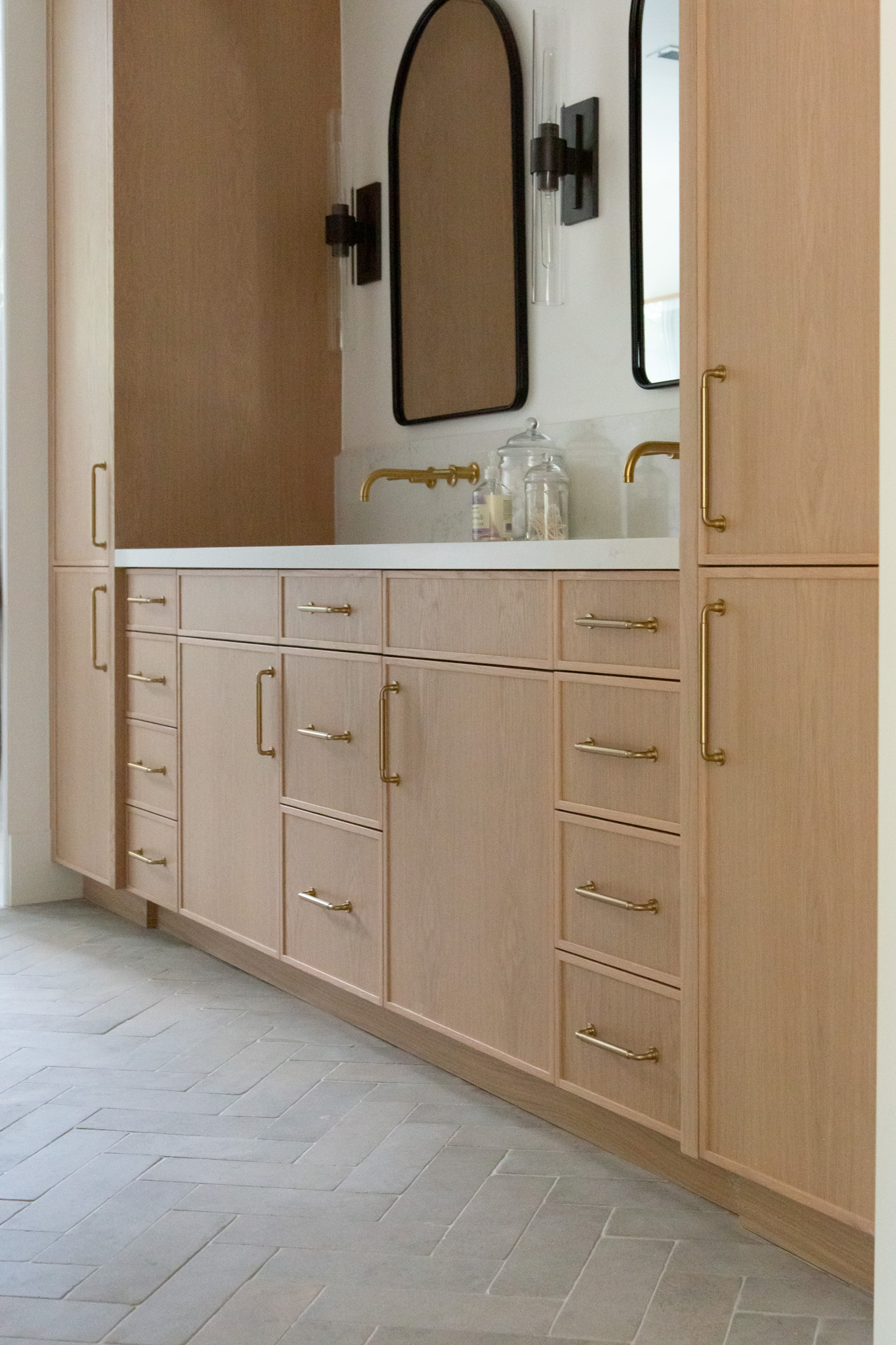 Light wood bathroom vanity with gold hardware, double faucets, a white countertop, two large mirrors, and built-in cabinets, set against a neutral-toned tiled floor.