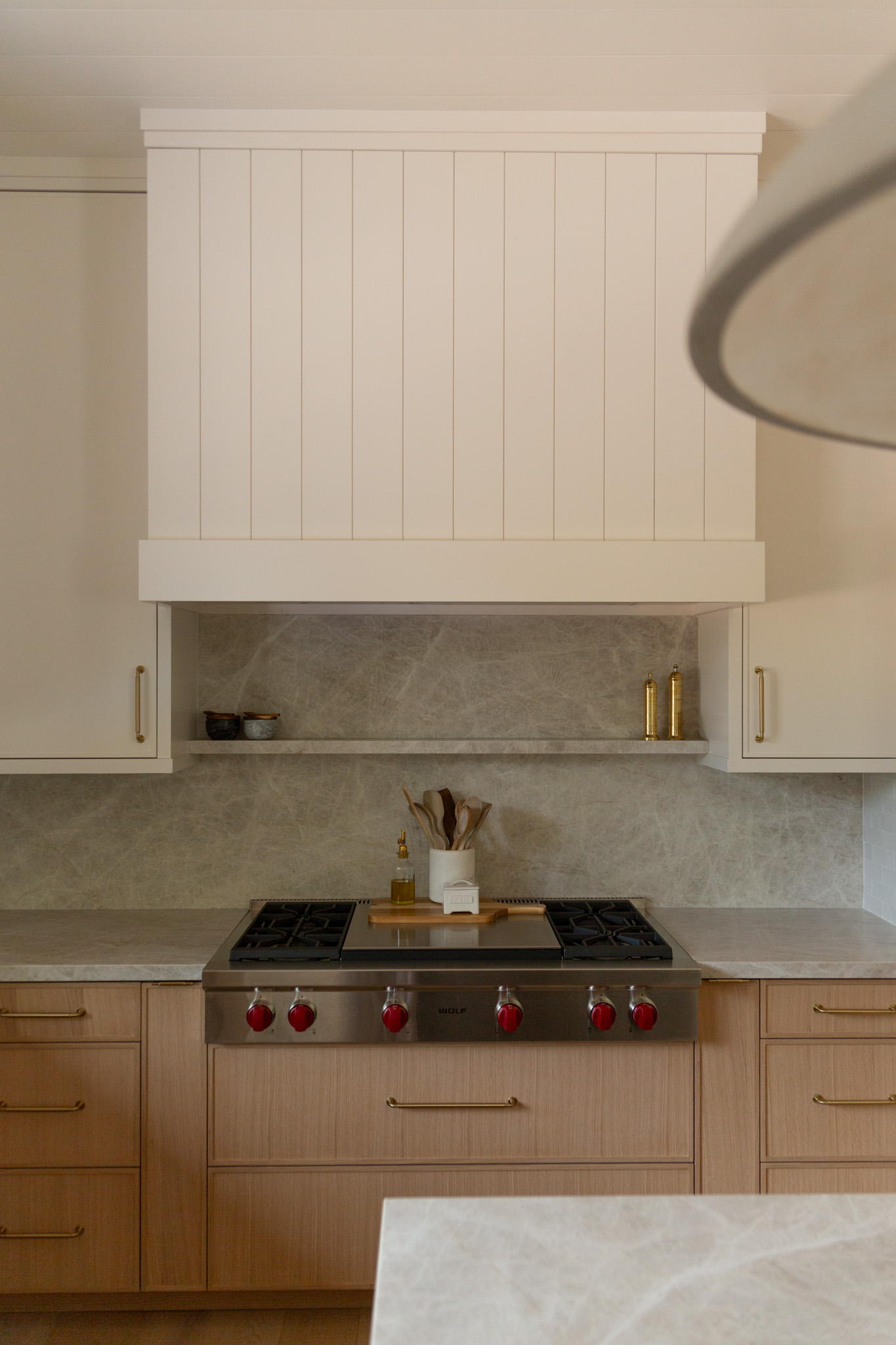 A modern kitchen with a stainless steel stove featuring red knobs, light wood cabinets, a marble backsplash, and white cabinetry above.