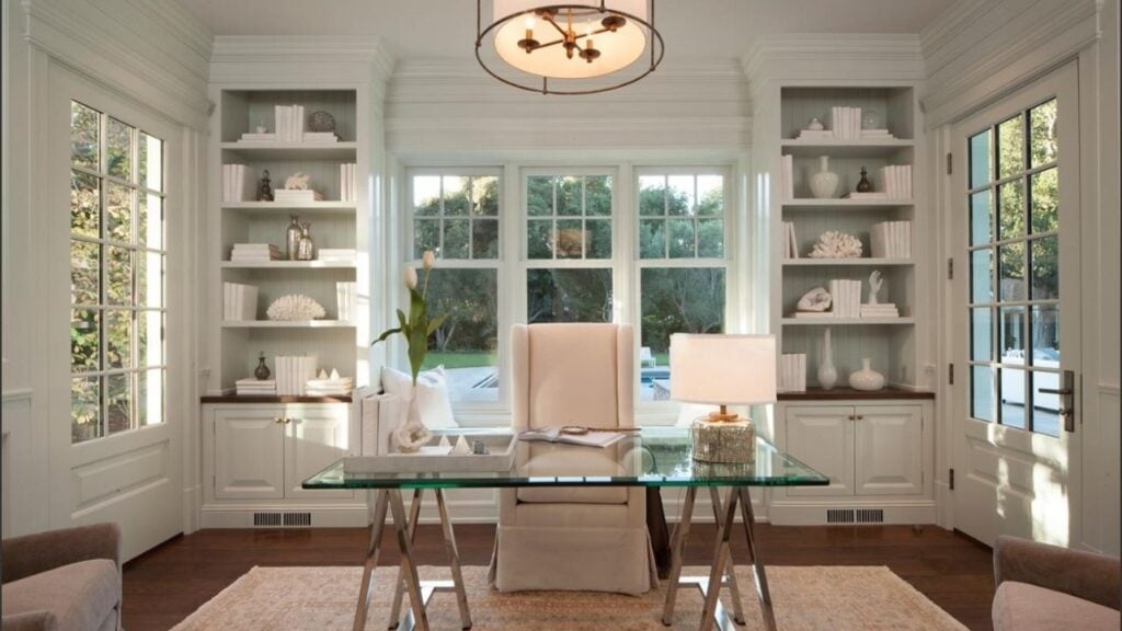 A home office featuring a glass desk with a white upholstered chair, centered in front of a window flanked by custom shelving in this home interior design.