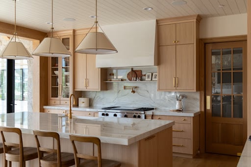 Modern kitchen with light wood cabinets, marble countertops, a large island with barstools, pendant lights, a gas stove, and glass door pantry—showcasing both style and excellent kitchen cabinetry cost value.