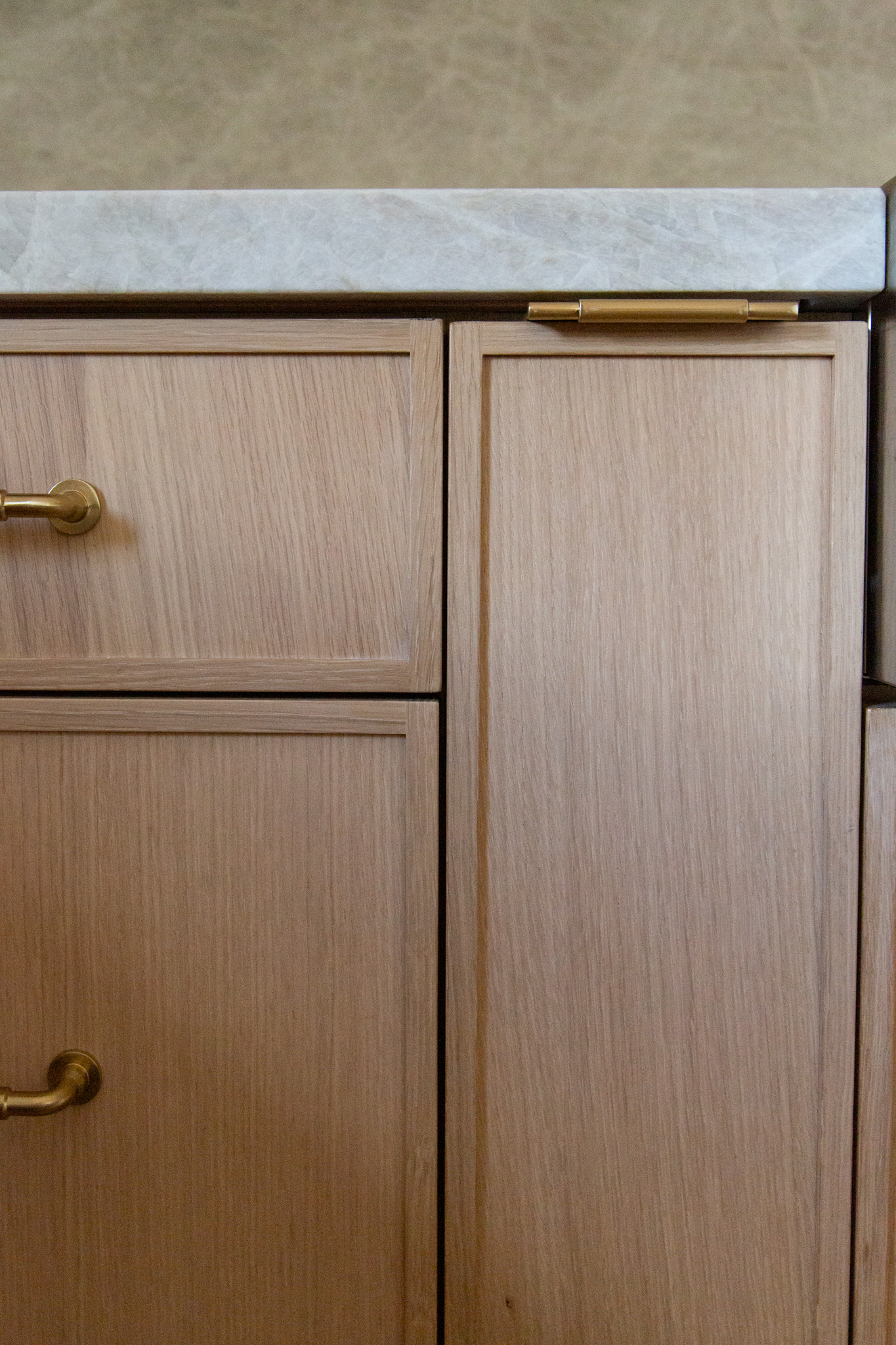 Light wood kitchen cabinets with brass handles and a marble countertop are shown in close-up.