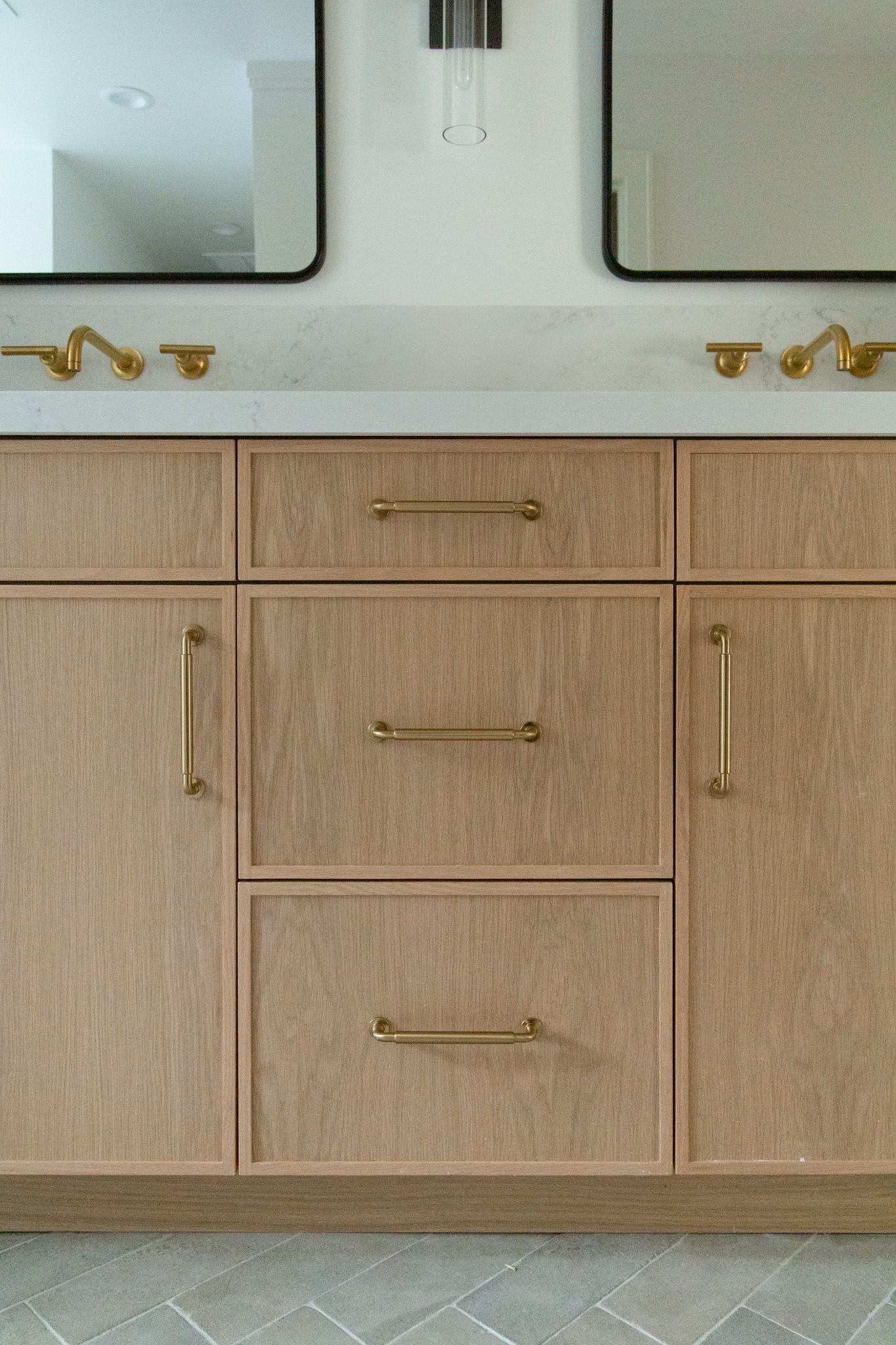 Light wood bathroom vanity with gold handles, white countertop, two square mirrors, and two gold faucets, set against a neutral wall and gray herringbone tile floor.