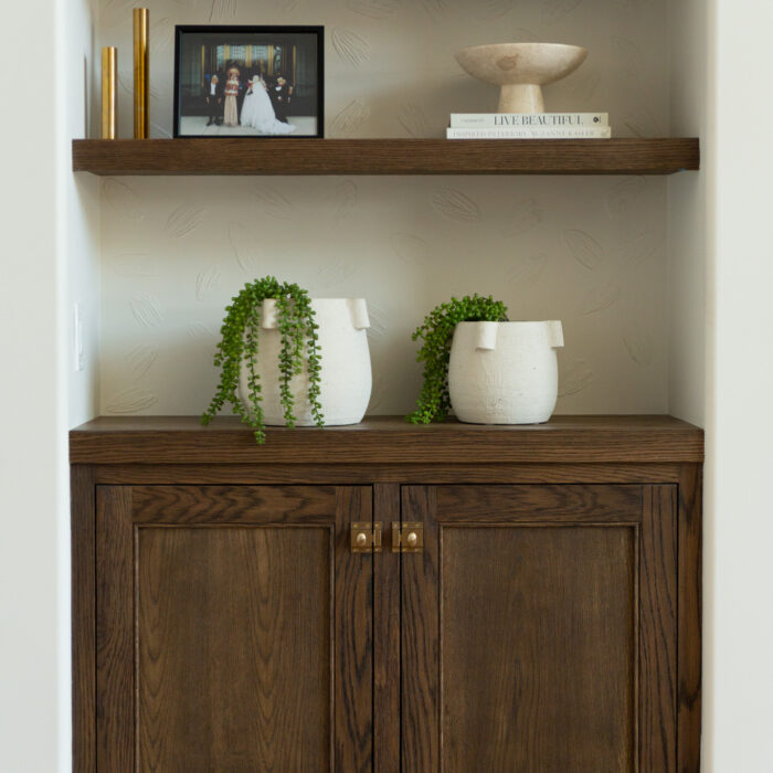 Two wooden shelves above a dark wood cabinet display decor items, including plants, vases, books, a bowl, a framed photo, and a gold decorative object.