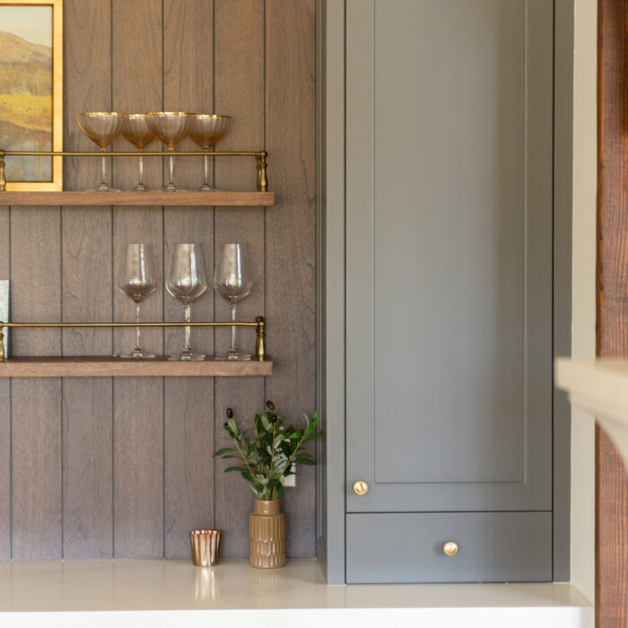 A modern kitchen bar area with gray cabinets, gold handles, glass shelves holding drinkware, a small plant in a gold pot, and a built-in wine fridge.