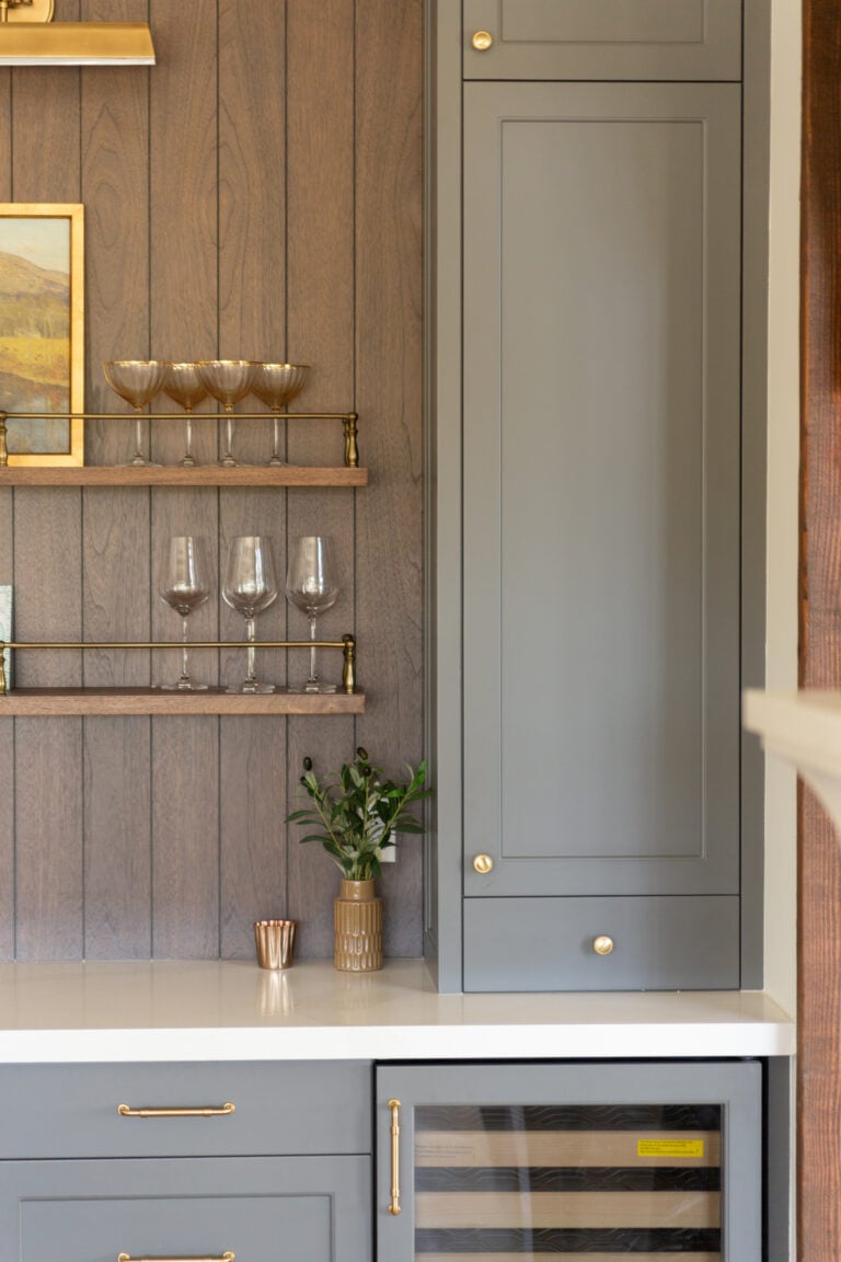 A section of a modern kitchen featuring gray cabinets, a small wine fridge, glassware on shelves, a potted plant, and a white countertop.