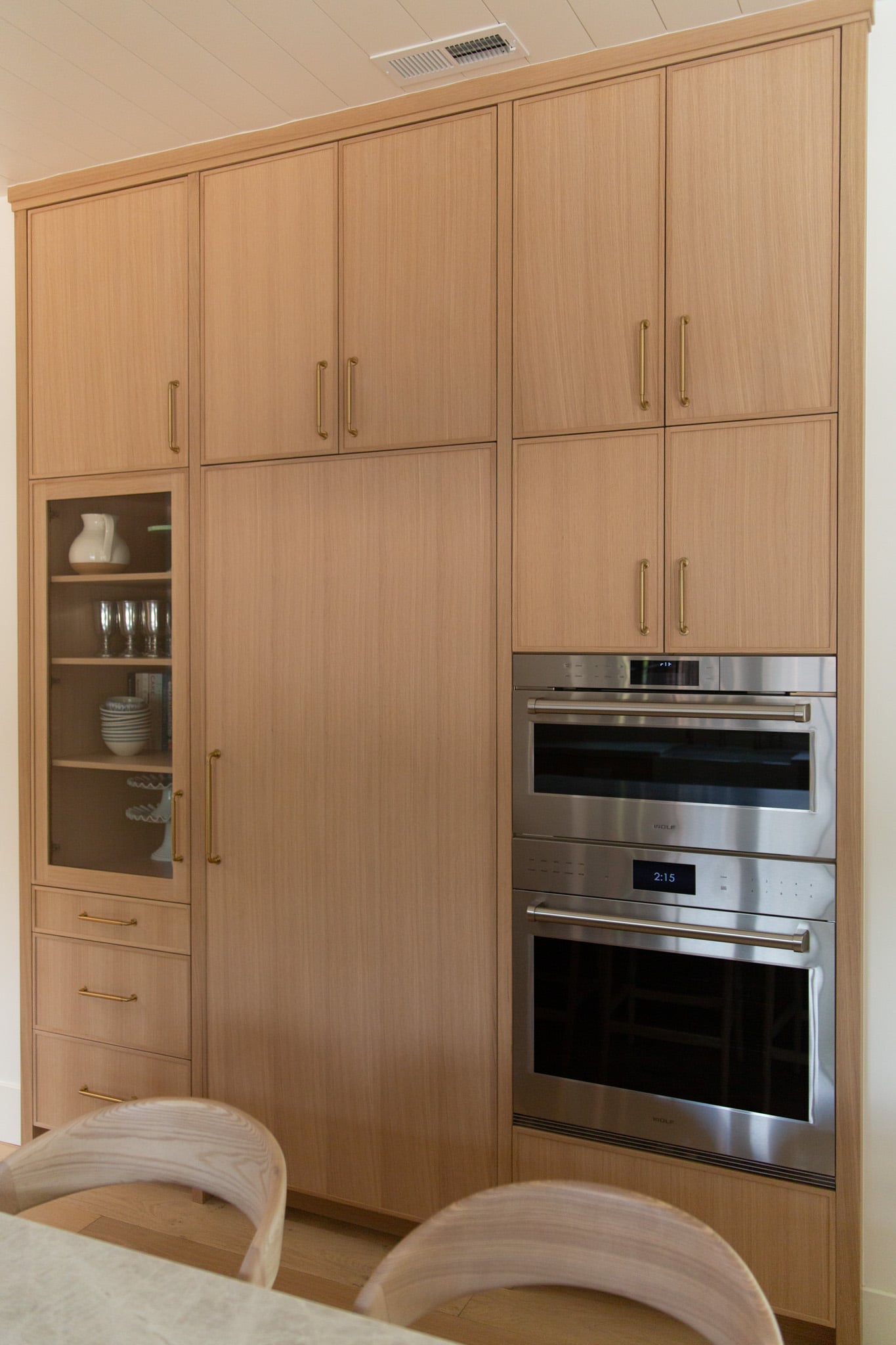 Built-in wooden kitchen cabinets with gold handles, glass-front display shelves, and two stainless steel wall ovens. A table and chairs are partially visible in the foreground.