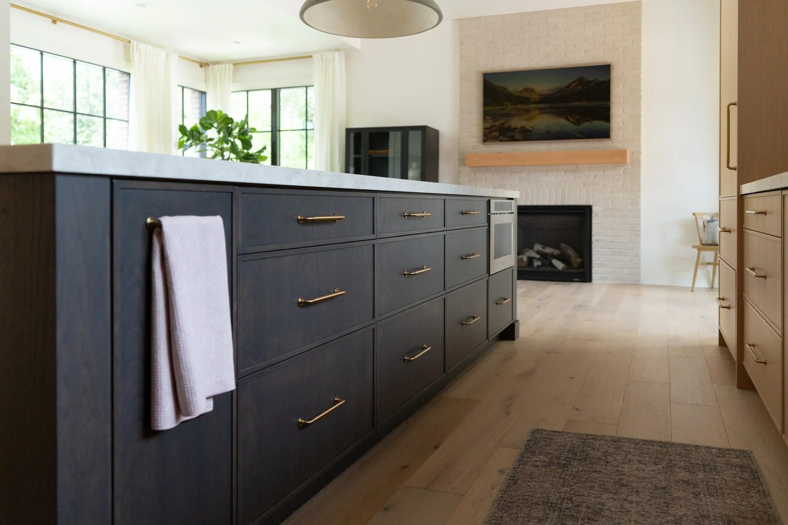 A modern kitchen with dark cabinets, gold handles, a towel hanging on the island, light wood floors, a fireplace, and a landscape painting above the mantle.