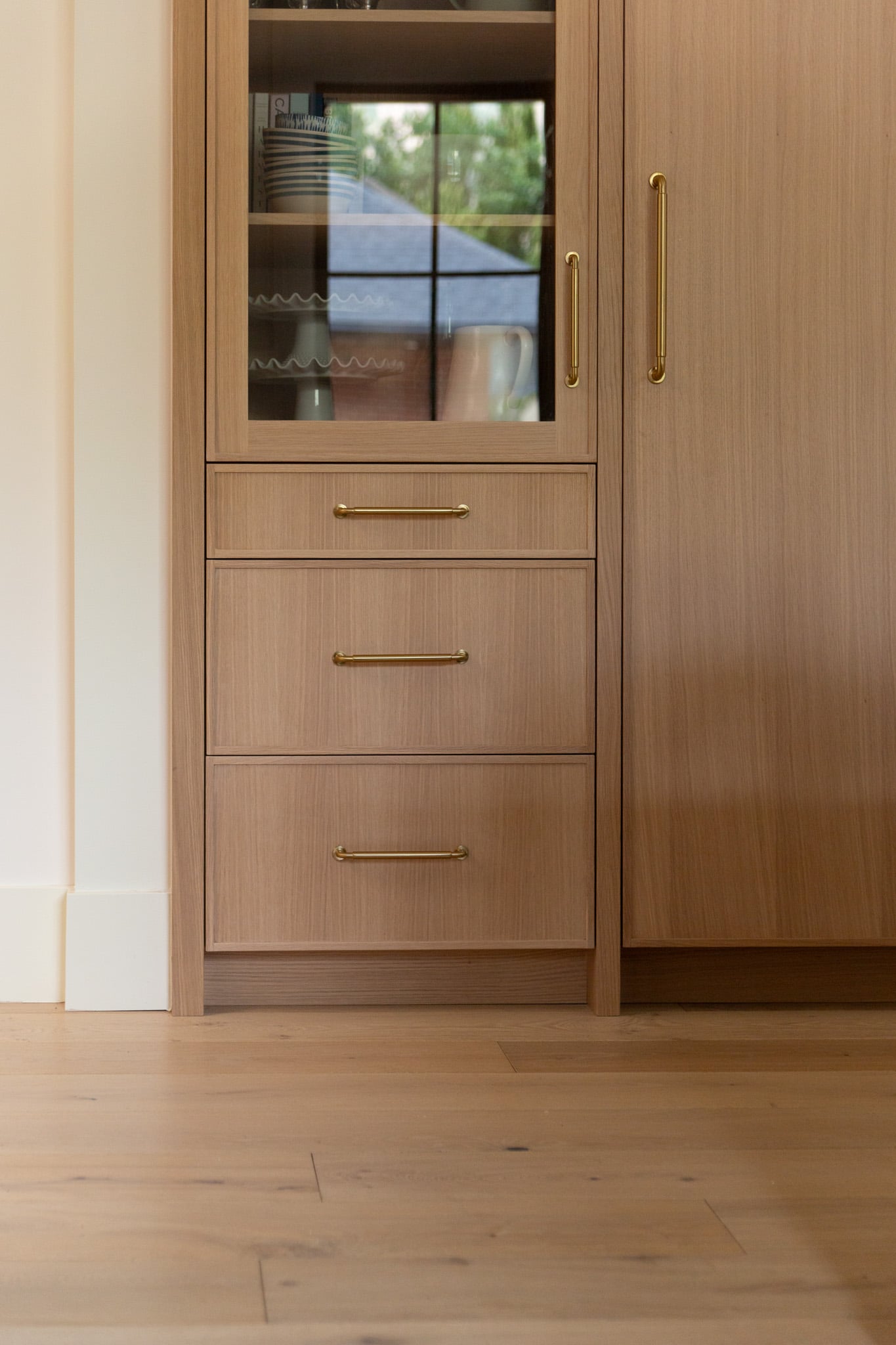 Light wood cabinet with three drawers and gold handles, glass-fronted upper section reflecting a window, and adjacent tall cupboard, set on a light wood floor.