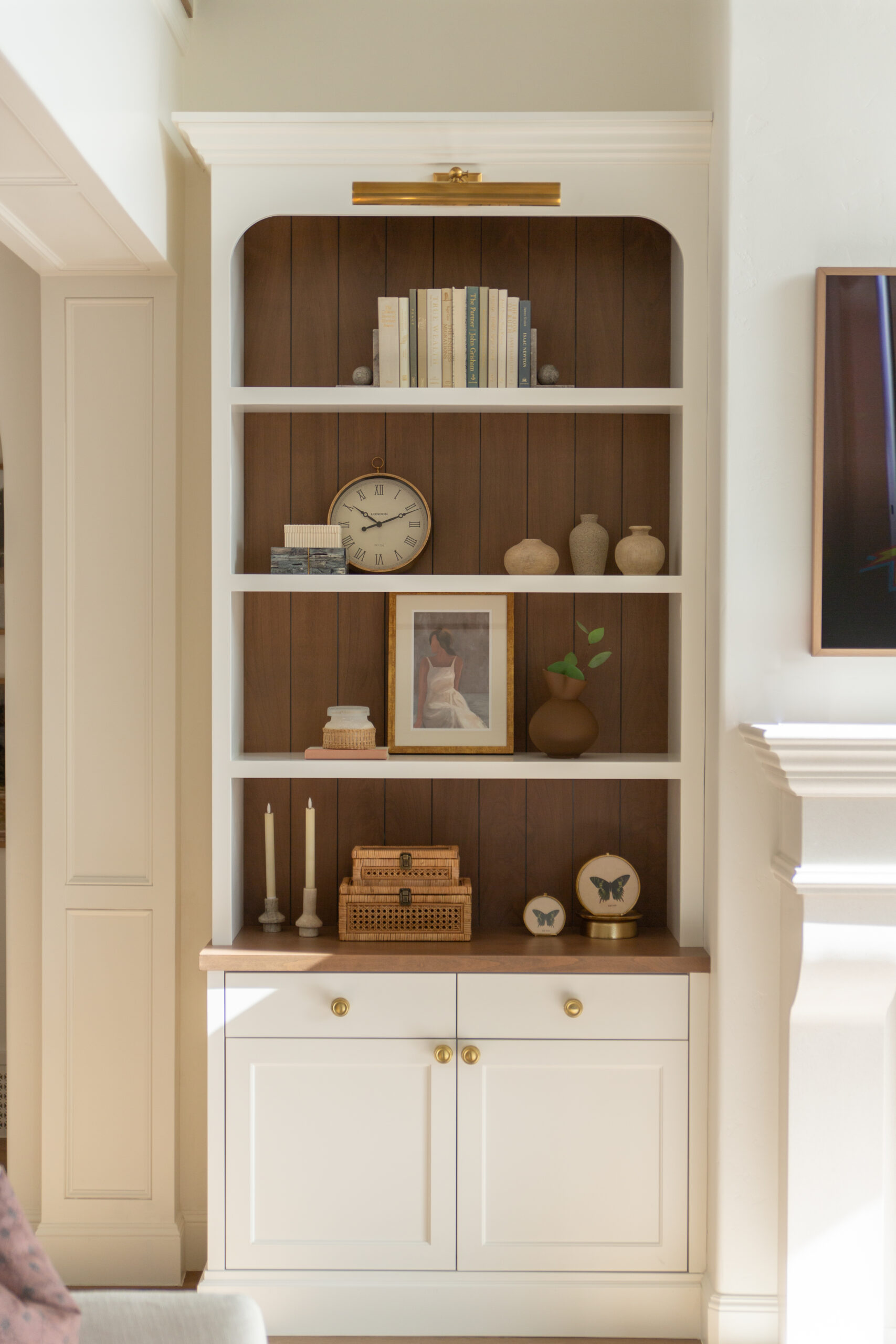 Custom Furniture Description: White custom built-ins form a stunning bookshelf, thoughtfully arranged with books, a clock, framed photo, vases, candles, wicker baskets, and decorative objects—including butterfly art—displayed neatly on the wooden shelves. This piece showcases exceptional craftsmanship and custom furniture design perfect for any Utah home.