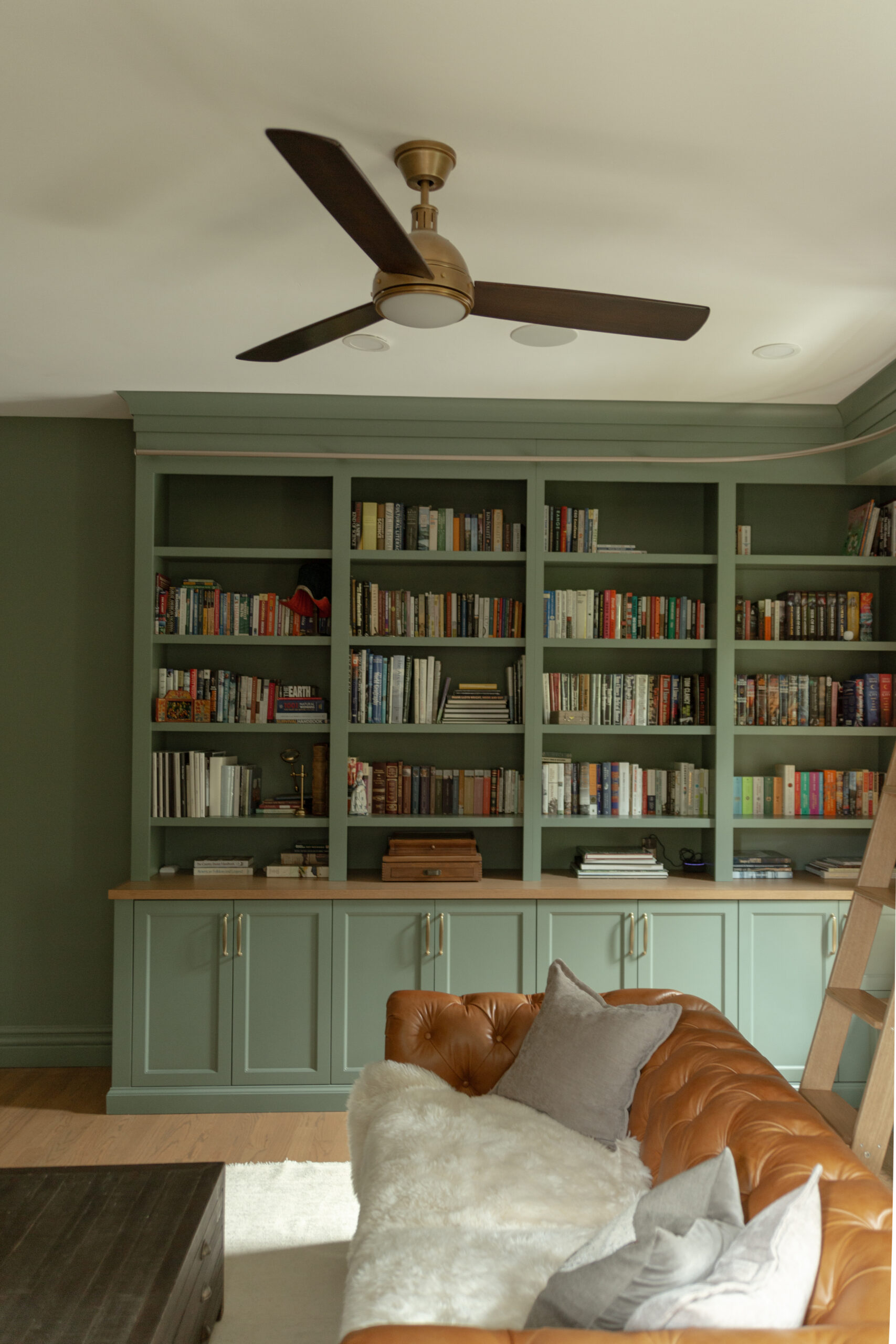 A green custom built-in bookshelf filled with books stands behind a brown leather sofa with pillows and a fur throw, under a ceiling fan—perfectly blending cozy style with the quality craftsmanship you’ll find in many Utah homes.