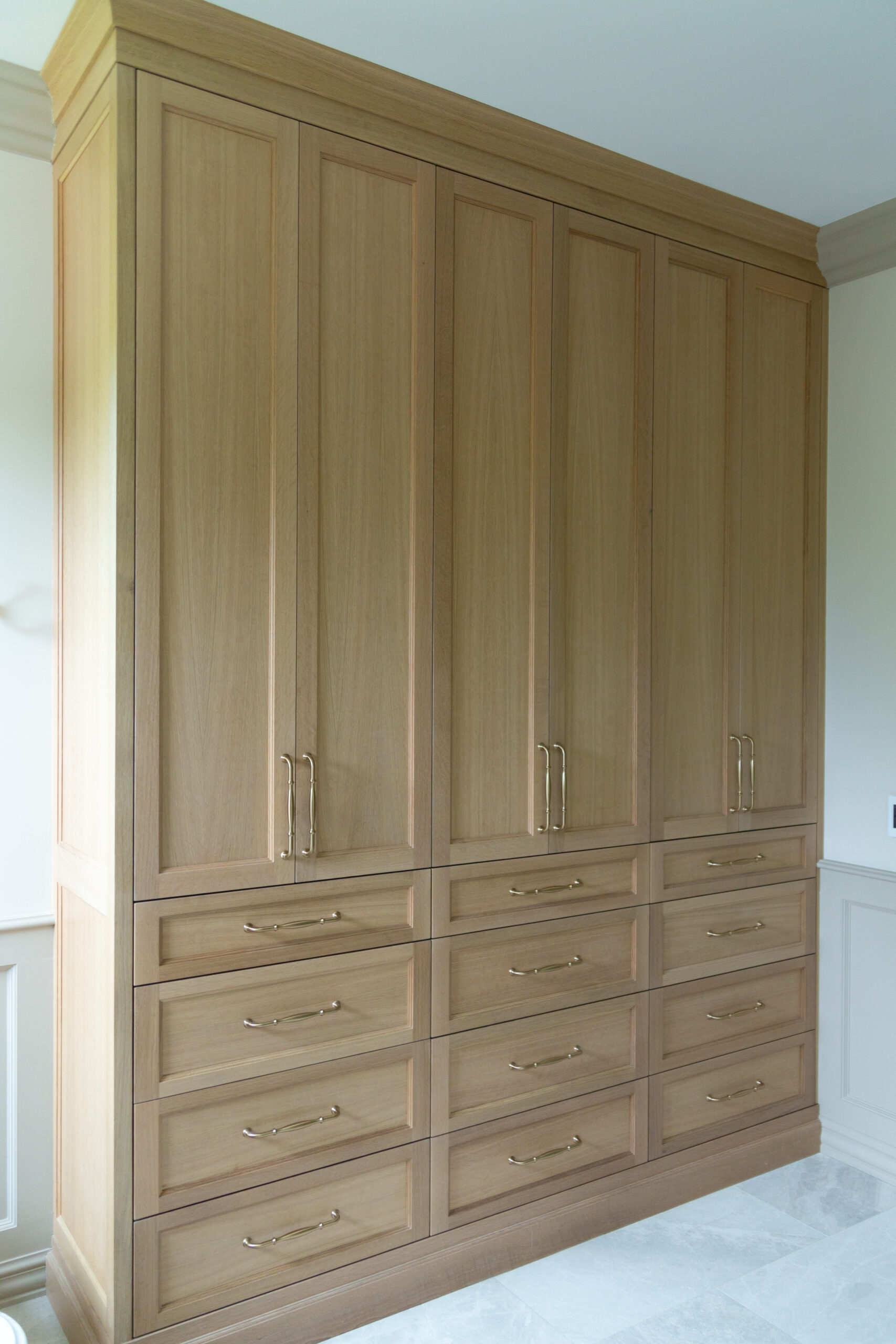Tall wooden wardrobe with six doors and twelve drawers, featuring brass handles, placed against a light-colored wall in a bright room—an elegant alternative to traditional garage cabinetry for those seeking refined storage solutions in Utah.