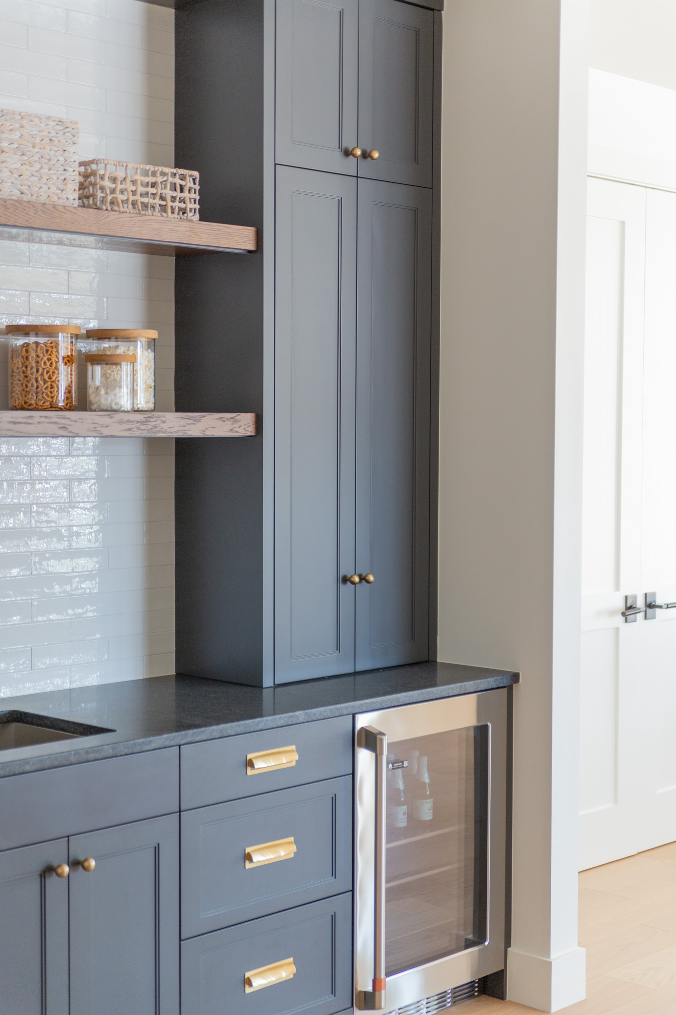 Modern kitchen corner with dark blue cabinets, gold handles, open wooden shelves, glass storage jars, and a built-in stainless steel mini fridge next to a white tiled wall—an elegant showcase of Custom Cabinets Utah perfect for any contemporary home.