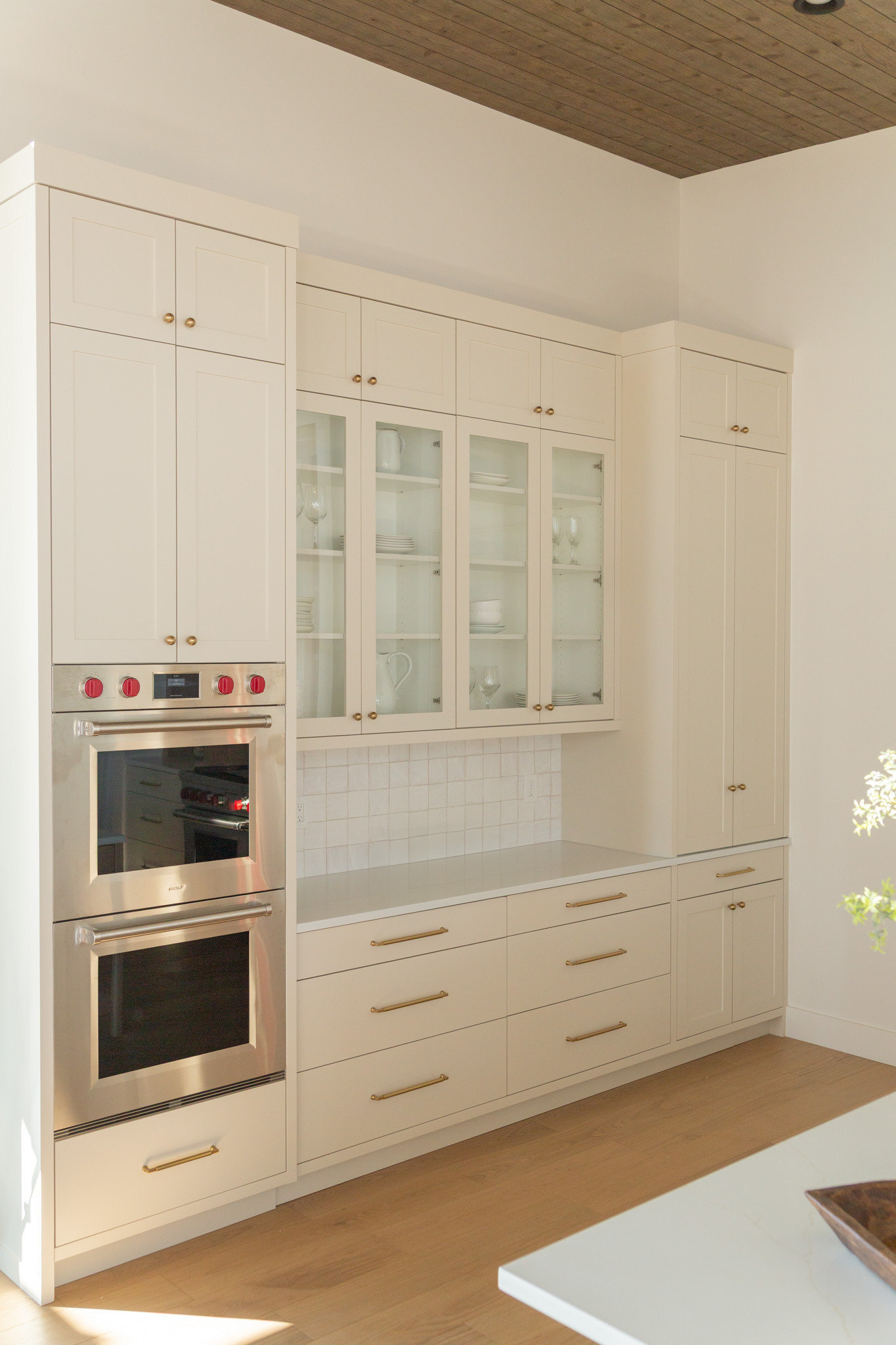 Bright modern kitchen with cream cabinets, glass-front upper doors, custom built-ins, built-in double oven, gold hardware, and wood flooring. White tiled backsplash and dishes are visible in the cabinets—perfect for showcasing Utah-inspired design and craftsmanship.