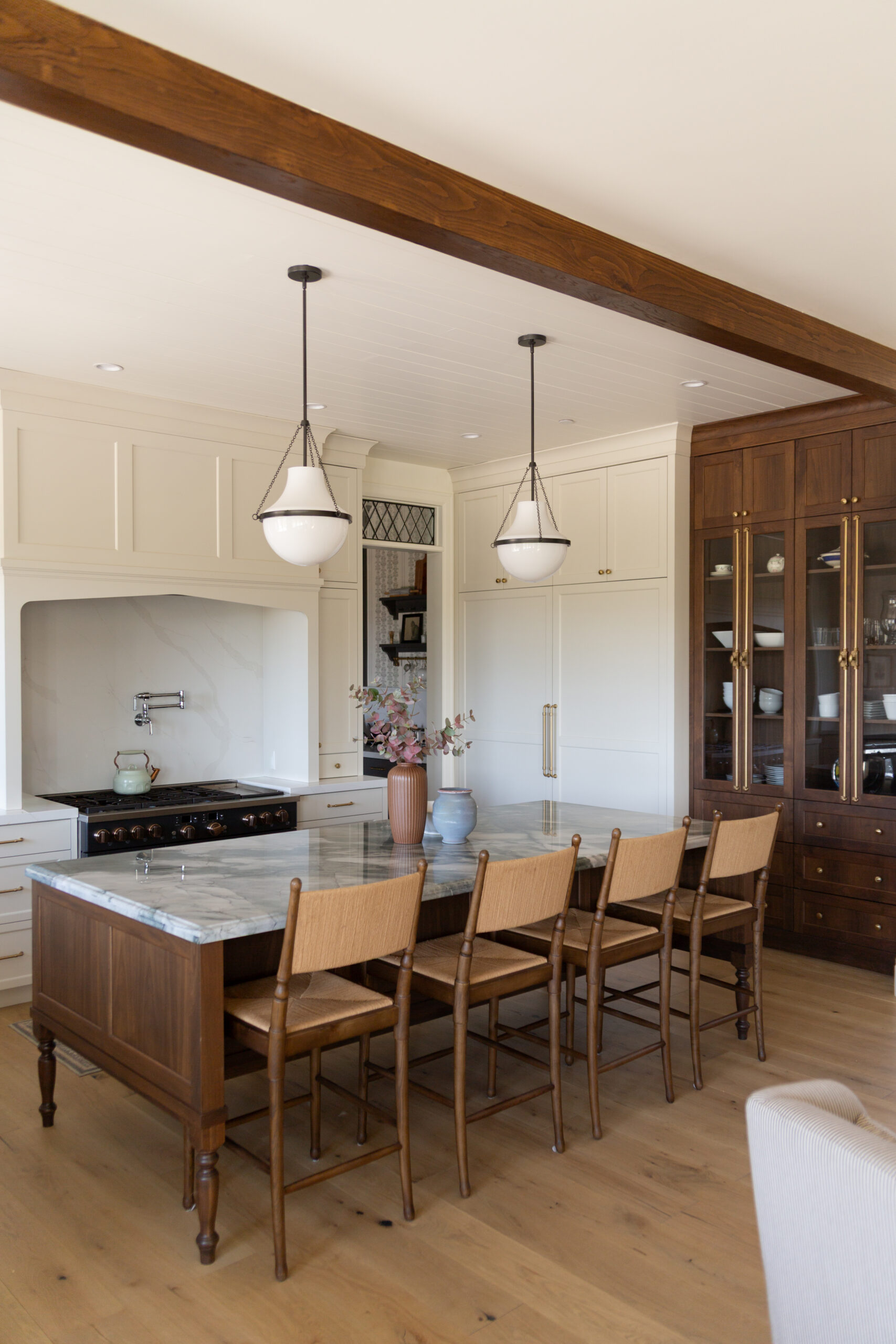 A modern kitchen with a marble island, five wooden chairs, pendant lights, white cabinetry, and a dark wood oven range. A vase with flowers sits on the island.