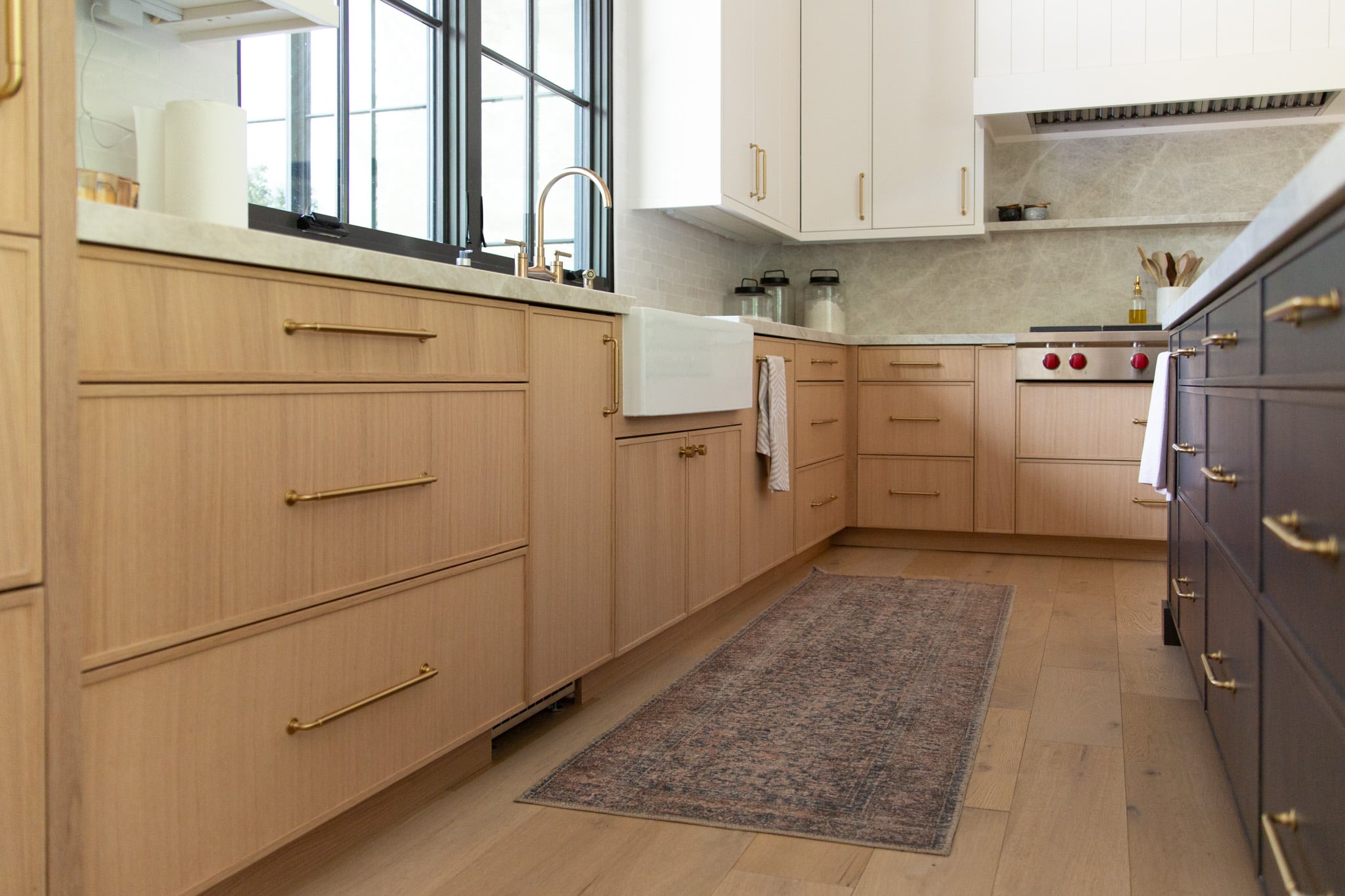 Modern kitchen with light wood cabinets, white countertops, brass handles, a farmhouse sink, an area rug, and large windows letting in natural light.