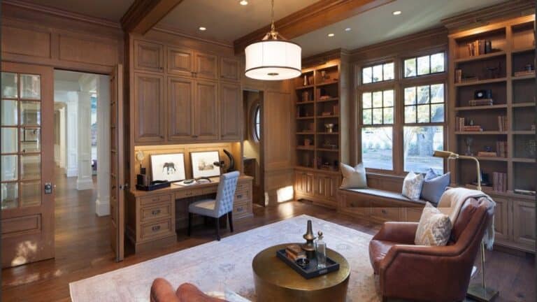 Home office with wood-paneled walls, a built-in desk, floor-to-ceiling bookshelves featuring modern home shelving, a window seat, and leather armchairs around a circular coffee table.
