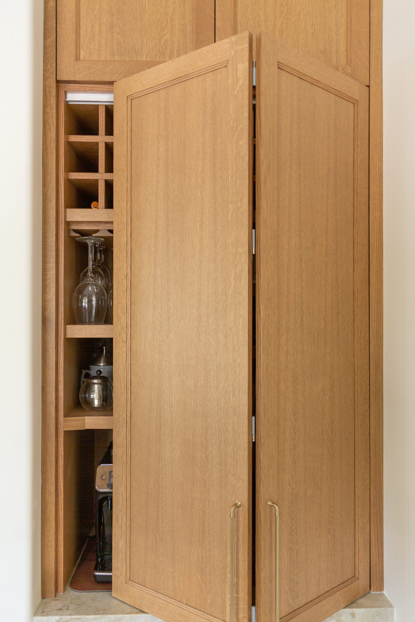 A wooden cabinet with open doors reveals shelves storing wine bottles, glassware, a coffee maker, and a kettle.
