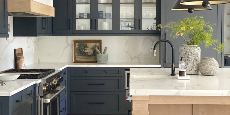 Modern kitchen with navy cabinets—a standout in today’s kitchen cabinetry trends—light wood island, marble countertops, gold accents, glass-front cabinets, black faucet, and decorative vases with greenery on the island showcase a fresh take on kitchen cabinet design.