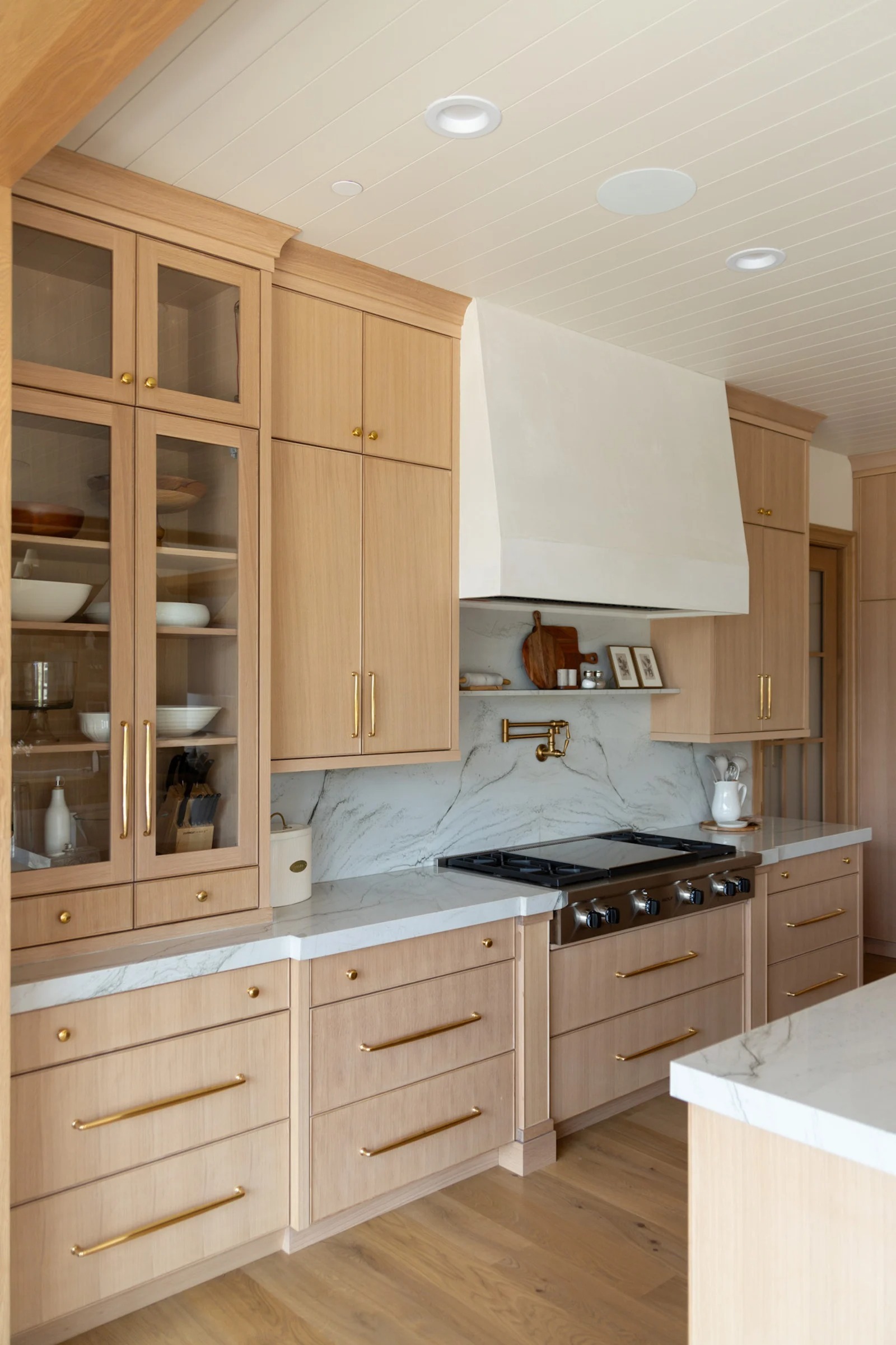 Modern kitchen with light wood cabinets, marble countertops, brass hardware, a gas stove, and a white range hood. Custom pantry cabinets add tailored storage solutions, while shelves display dishes and accessories. Natural light brightens the space, making it an inviting Utah home kitchen.