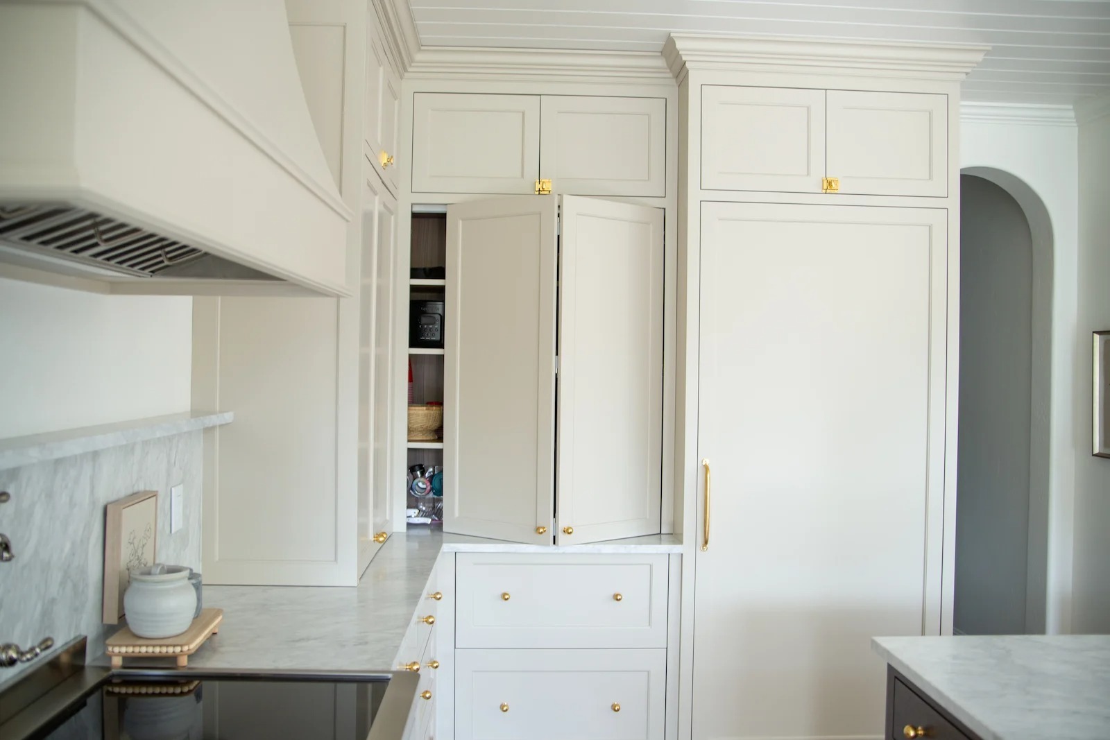 A modern white kitchen with gold hardware, a marble countertop, and a tall corner cabinet with bi-fold doors partially open to reveal shelves and stored items inside—an elegant space that showcases the convenience and style of custom pantry cabinets.
