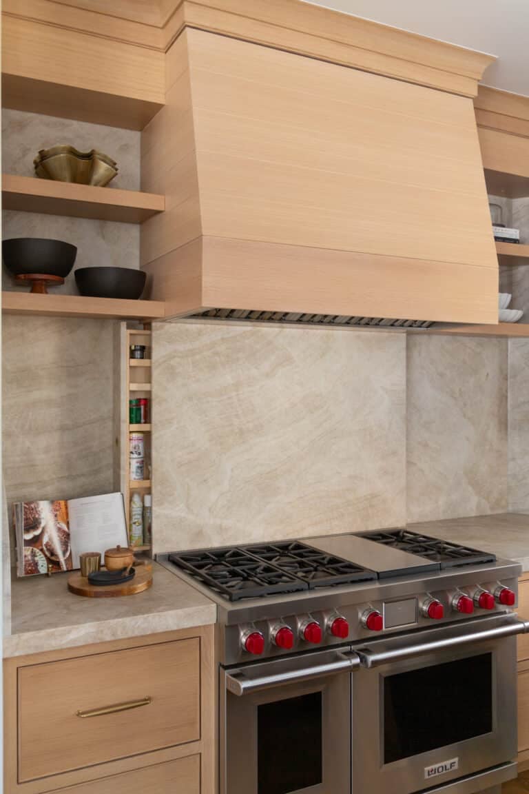 Modern kitchen with a stainless steel gas stove, red knobs, wooden cabinetry crafted by a renowned cabinet company, open shelves with bowls, and a marble backsplash. Cookbooks and spices are arranged on the counter.