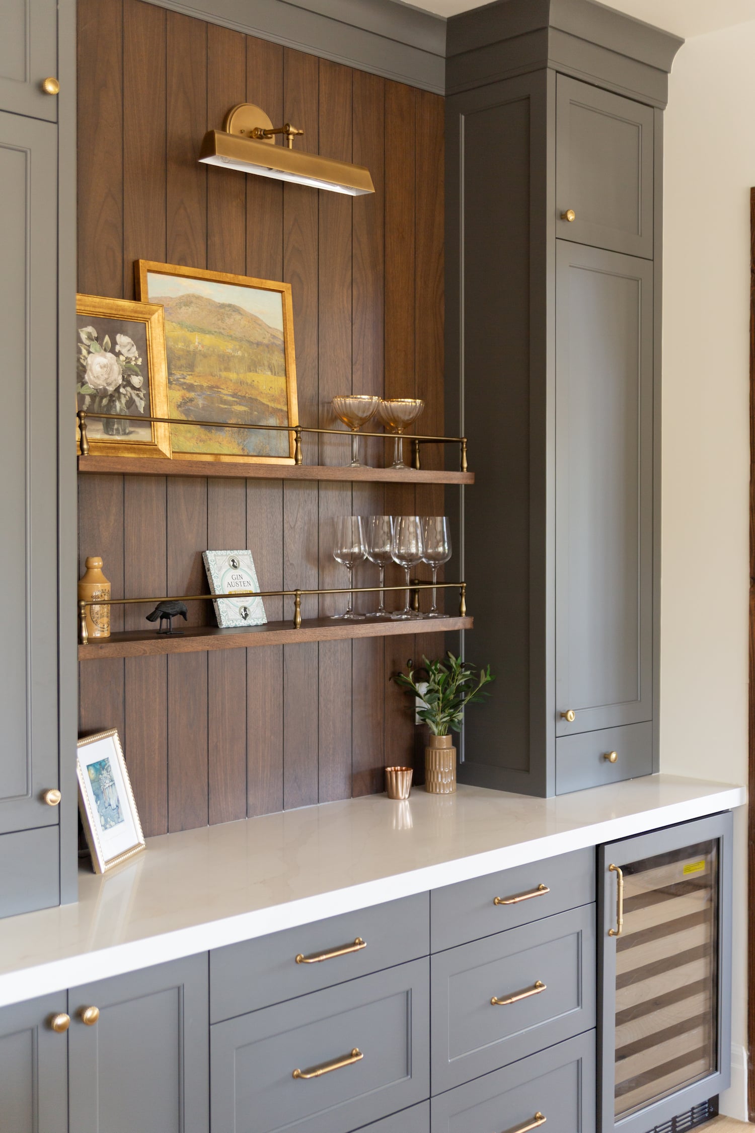 Modern home bar area with gray cabinets, gold hardware, a built-in wine fridge, wood panel backsplash, glass shelves, and decorative artwork and glasses on display.